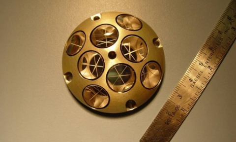 The device is a gold-colored ball with a large square flat plate running through the middle and out the sides of the sphere. The gold-colored sphere is hollow with many holes going through it. These holes have a black edge as well as are equidistant from one another.