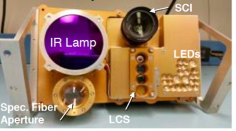 A golden device, known as LETS, sits on a white desk. The IR lamp shines a dark purple while the SCI is a dark black. Most of the devices attached to LETS have a circular shape including: the IR lamp, SCI, Spec. Fiber Aperture, LCS, and the LEDs. 