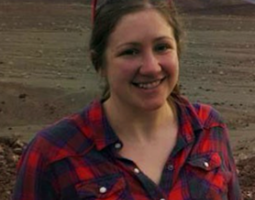Photo of a smiling woman with sunglasses on top of her head, wearing a red and black plaid shirt and standing in a field.